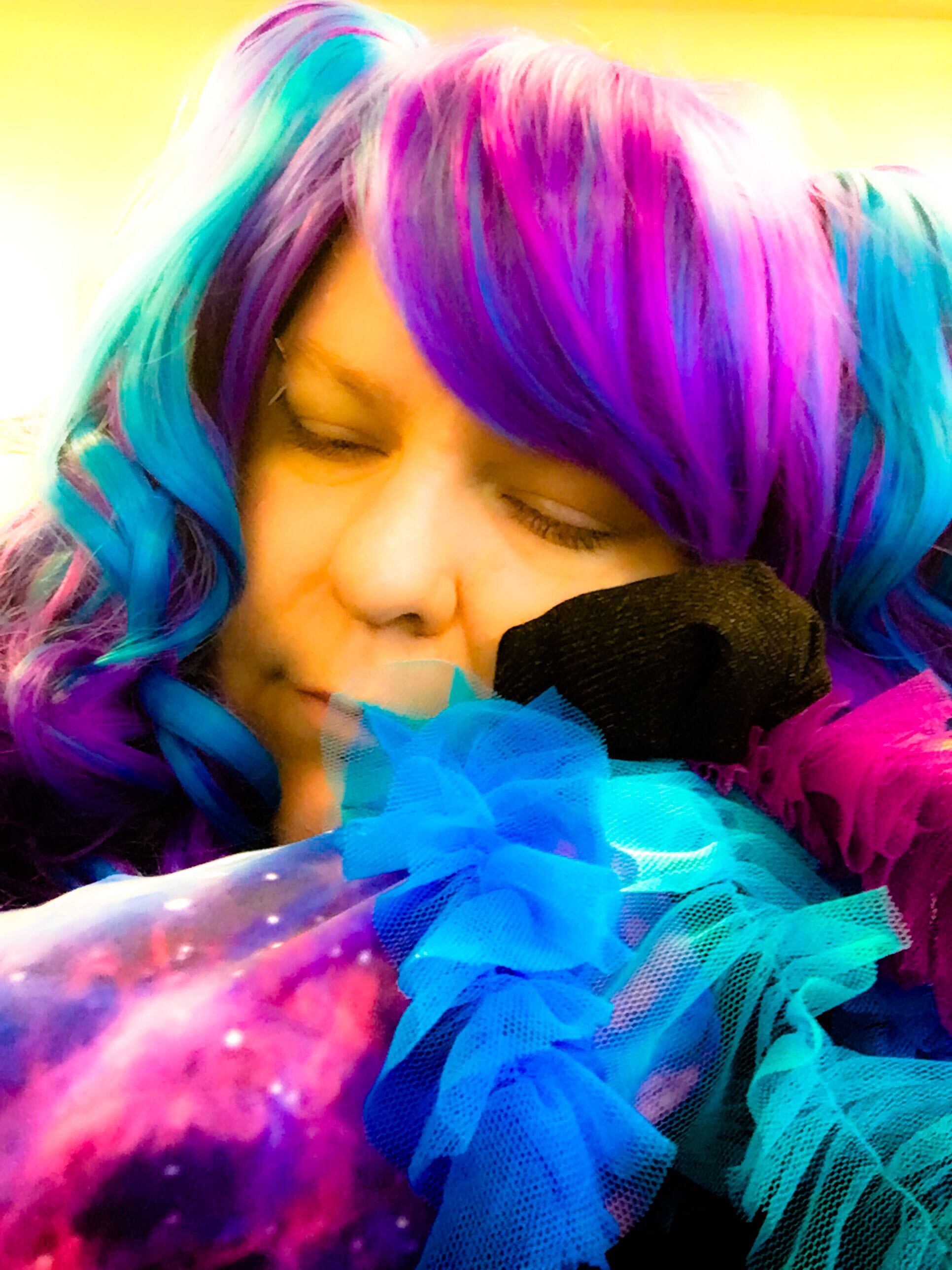 A person in a long blue and purple and pink wig resting their head on their arms, eyes closed. Their sleeves are space-patterned and a small home-made stuffed jellyfish half conceals their face.